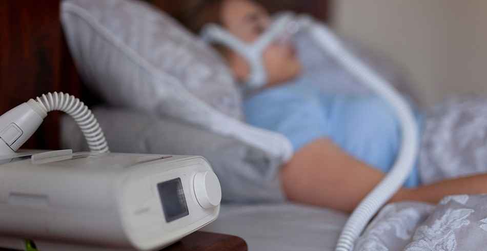 woman wears CPAP mask connected to machine beside bed (life support)
