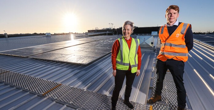 Smiling woman and man in high-vis vests on top of Coles supermarket roof with solar panels in the background (Origin)