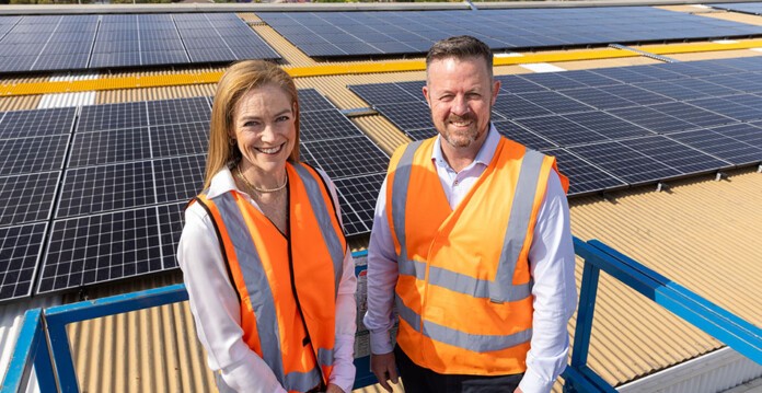 Smiling woman and man in hi-vis orange vests stand in front of massive rooftop solar installation (detmold)