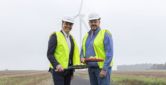 Two men in hard hats and high-vis vests hold tennis racquets with wind turbine in the background (pacific blue)